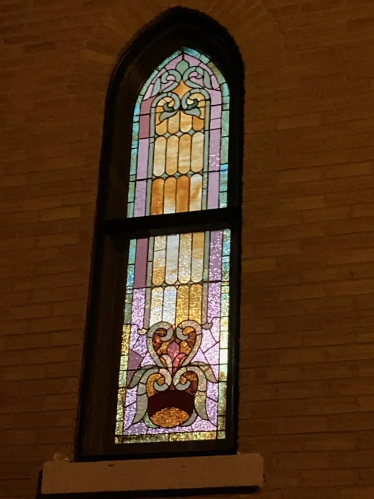 The tall arched west stained-glass window in pink, amber and green glass set in the brick wall